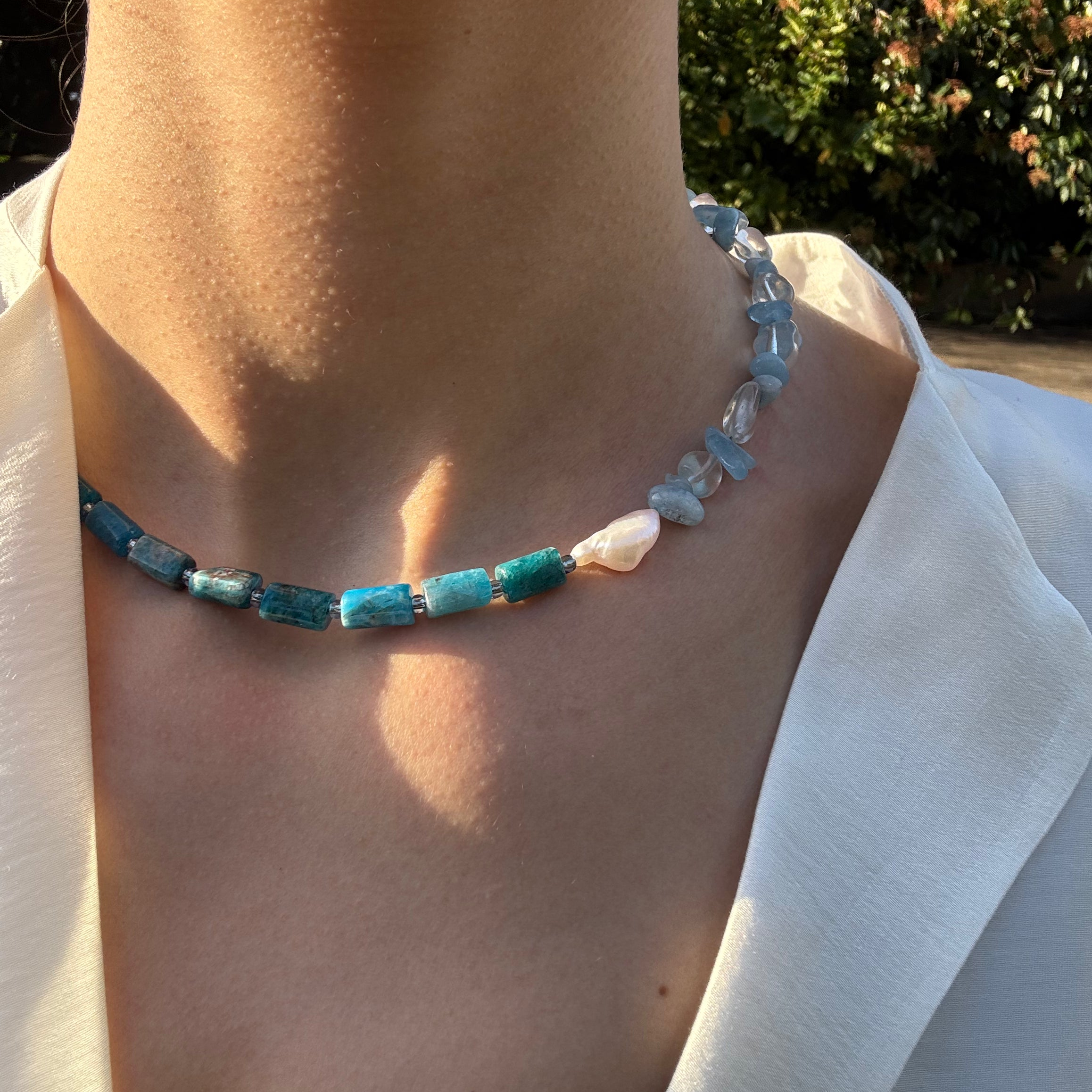 A close-up of a person's neck and chest wearing a beaded necklace made of turquoise-blue rectangular beads, light blue irregular beads, and a few white beads, styled with a white top outdoors.